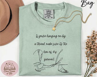 May include: A pale green Comfort Colors t-shirt with the text "If you're hanging on by a thread, make sure its the hem of His garment." A gold star pendant necklace is on the shirt. The shirt has a graphic and the text "Matthew 9:20-21".