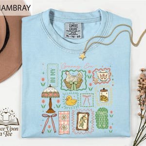 a light blue t-shirt with a colorful graphic design, including a lamp, teapot, flowers, and a bird, along with a gold chain and a star pendant.