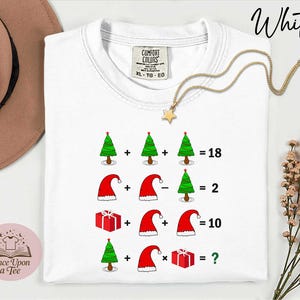 May include: White t-shirt featuring a Christmas-themed mathematical equation. The equation uses images of Christmas trees, Santa hats, and gift boxes. The text "Once Upon a Tee" is printed on a pink circle. The shirt has a Comfort Colors tag.