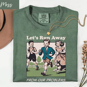 May include: A moss green t-shirt featuring a graphic print of two children and a dog running in a field. The text "Let's Run Away" appears above the illustration, and "FROM OUR PROBLEMS" is printed below. The shirt has a Comfort Colors label.
