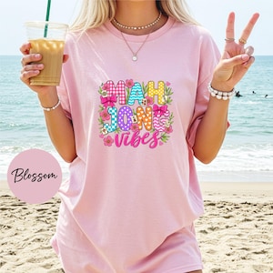 May include: A light pink t-shirt featuring a colorful graphic print that reads Mahjong Vibes in bold, patterned letters. The design is decorated with pink flowers, green leaves, small pink bows, and miniature mahjong tiles in a vibrant style.