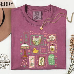 a purple t-shirt with a variety of colorful illustrations and text, including a bird, flowers, a teapot, and other decorative elements. The t-shirt is displayed on a white background, and there is a hat and some flowers nearby. The text "BERRY" is visible, and the phrase "Once Upon a Tee" is also present.