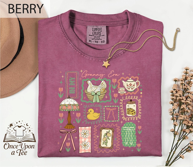 a purple t-shirt with a variety of colorful illustrations and text, including a bird, flowers, a teapot, and other decorative elements. The t-shirt is displayed on a white background, and there is a hat and some flowers nearby. The text "BERRY" is visible, and the phrase "Once Upon a Tee" is also present.