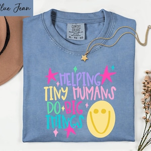 May include: A blue jean colored t-shirt with a graphic print that reads HELPING TINY HUMANS DO BIG THINGS in multi-colored lettering. The design includes a yellow smiley face and pink stars. The shirt has a Comfort Colors brand neck label.