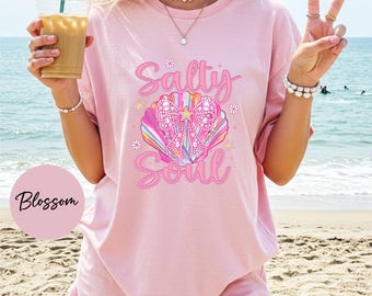 Può includere: Una t-shirt a maniche corte rosa chiaro con la grafica di una conchiglia colorata e un grande fiocco a pois. Il testo Salty Soul è stampato in corsivo rosa sopra e sotto la conchiglia, circondato da piccole icone di fiori e stelle.