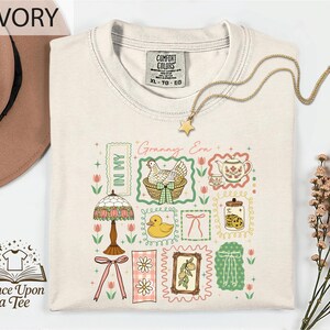 a white t-shirt with a colorful graphic design, including illustrations of various objects such as a lamp, teapot, flowers, and a bird. The t-shirt is displayed on a white background, and there is a hat and some flowers nearby.