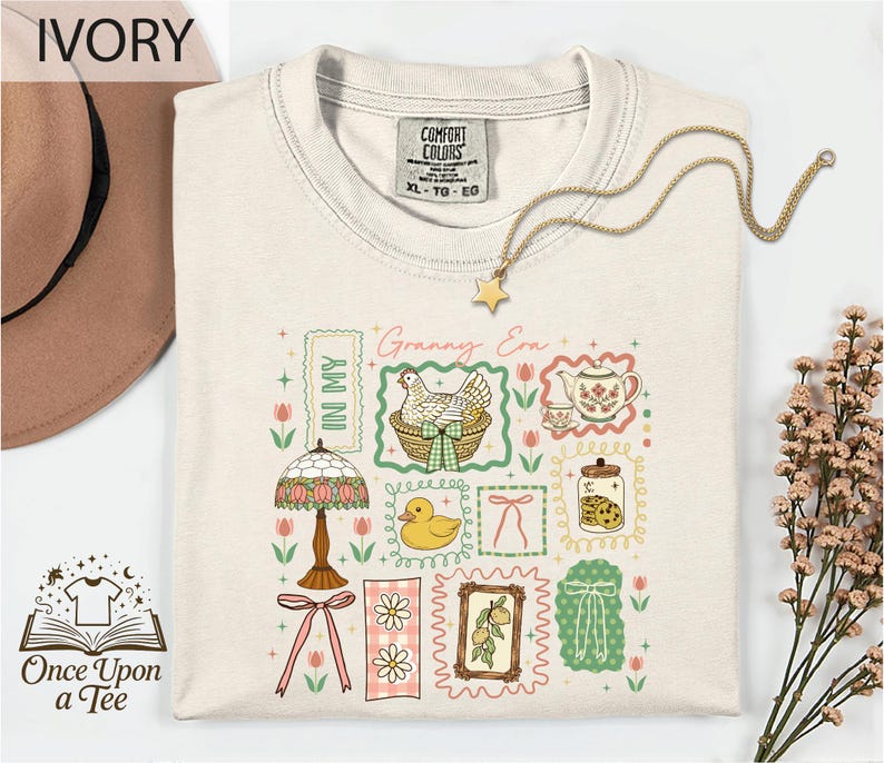 a white t-shirt with a colorful graphic design, including illustrations of various objects such as a lamp, teapot, flowers, and a bird. The t-shirt is displayed on a white background, and there is a hat and some flowers nearby.