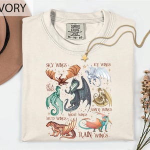 a white t-shirt with a graphic design of various dragon illustrations, including a red dragon, a green dragon, and a blue dragon, along with the text "Sky Wings", "Ice Wings", "Sea Wings", "Mud Wings", "Night Wings", "Rain Wings", and "Ice Wings". The t-shirt is displayed on a white background, with a hat and some flowers visible in the background.