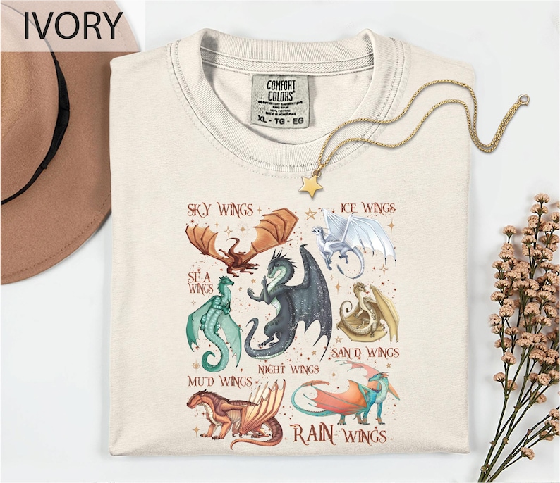 a white t-shirt with a graphic design of various dragon illustrations, including a red dragon, a green dragon, and a blue dragon, along with the text "Sky Wings", "Ice Wings", "Sea Wings", "Mud Wings", "Night Wings", "Rain Wings", and "Ice Wings". The t-shirt is displayed on a white background, with a hat and some flowers visible in the background.