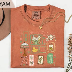 a red t-shirt with a colorful graphic design, including a bird, flowers, and other decorative elements. The t-shirt is folded and displayed on a white surface, with a hat and some flowers nearby.