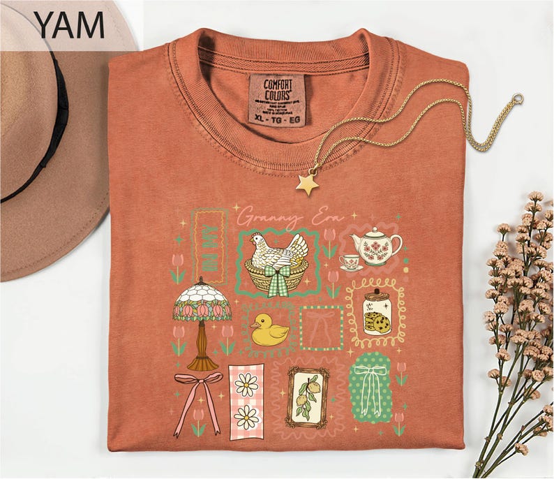 a red t-shirt with a colorful graphic design, including a bird, flowers, and other decorative elements. The t-shirt is folded and displayed on a white surface, with a hat and some flowers nearby.