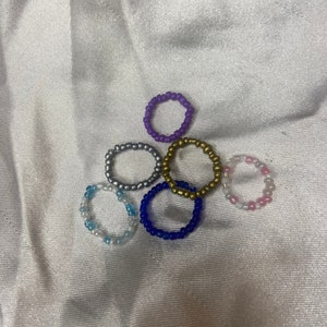May include: Six beaded rings in various colors, including purple, gold, silver, blue, and pink. The rings are made of small beads and are stacked on top of each other.