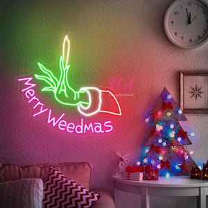 May include: A neon sign saying "Merry Weedmas" in pink, depicting a hand holding a cannabis plant. The sign is outlined in white, green, and red. A Christmas tree with lights and bows is on a table.