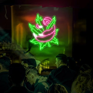 Pode incluir: Uma placa de n&eacute;on com um pregui&ccedil;a rosa enroscado dentro de uma folha verde. A placa est&aacute; pendurada em um bar ou restaurante com pessoas em p&eacute; na frente dela.