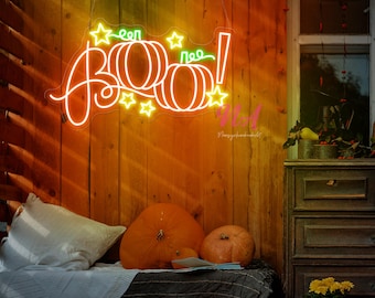 Op de afbeelding: Een neonbord met het woord "BOO!" in een speels lettertype, met pompoen- en stervormen. Het bord hangt tegen een houten muur, met pompoenen en een gezellige zithoek op de voorgrond. Het bord straalt een warme oranje gloed uit.