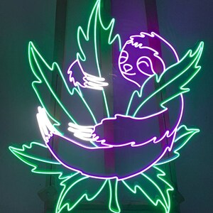 Pode incluir: Um letreiro de n&eacute;on que representa um bicho-pregui&ccedil;a roxo enroscado dentro de uma folha verde. O bicho-pregui&ccedil;a tem uma express&atilde;o feliz e est&aacute; rodeado de folhas verdes.