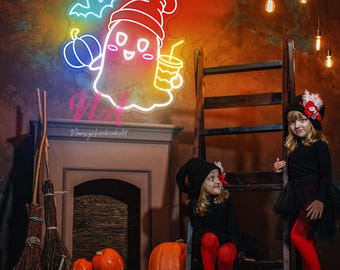 May include: Two children dressed in Halloween costumes stand in front of a fireplace with a neon sign of a ghost holding a drink. The sign is lit up in rainbow colors. The children are wearing black and red outfits and have on witch hats. There are pumpkins on the floor in front of them.