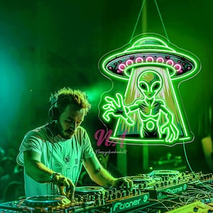 May include: A neon sign depicting a green alien emerging from a UFO, with pink and green accents. The sign is hanging above a DJ mixing music on turntables. The scene is illuminated with green light.