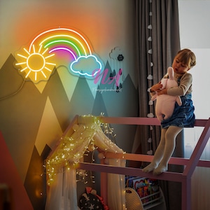 May include: A neon rainbow with a yellow sun and a blue cloud. The rainbow is lit up and is hanging on a wall with a pink and gray geometric pattern. There are fairy lights in the background.