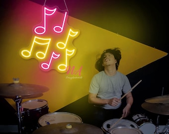 May include: A neon sign in the shape of musical notes, with pink and yellow illumination, hangs above a drum set. A person is playing the drums, with a yellow and black geometric background.