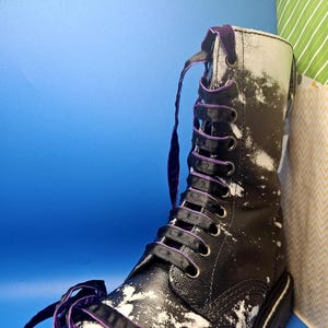 May include: Black leather boots with white paint splatter and purple ribbon laces. The laces are tied in a bow at the top of the boot.