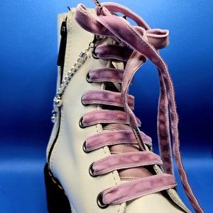 Może przedstawiać: Białe skórzane botki z zamkiem błyskawicznym z boku i sznurowadłami z fioletowego aksamitu. Sznurowadła są zawiązane w kokardę na górze buta.