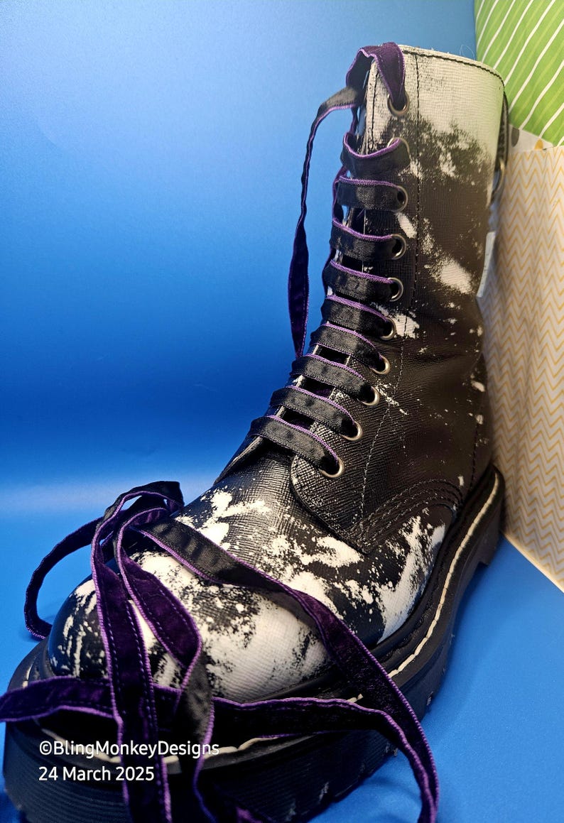 May include: Black leather boots with white paint splatter and purple ribbon laces. The laces are tied in a bow at the top of the boot.