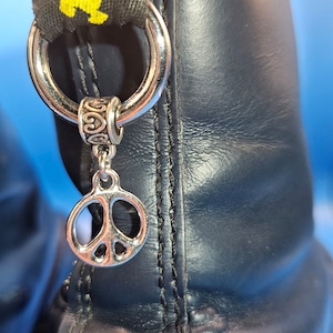 Könnte beinhalten: Nahaufnahme eines schwarzen Lederstiefels mit einem silbernen Peace-Zeichen-Anhänger. Der Anhänger hängt an einem silbernen Ring, der an der Zuglasche des Stiefels befestigt ist, mit gelbem Text auf schwarzem Hintergrund.