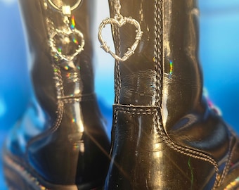 May include: Black patent leather boots with rainbow stitching and a barbed wire heart charm attached to a silver chain. The boots have a thick platform sole and a pull-on tab with the text "Dr. Martens" on it.