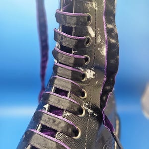 May include: Close-up of a black and white boot with purple laces. The boot has a white paint splatter design and a black ribbon trim.