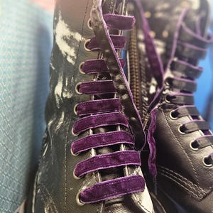 May include: A pair of black leather boots with purple velvet laces. The boots have a white and black tie-dye pattern on the upper part of the boot.