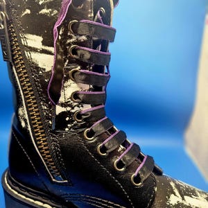 May include: Black leather boots with white paint splatter and purple laces. The boots have a side zipper and a white rubber sole.