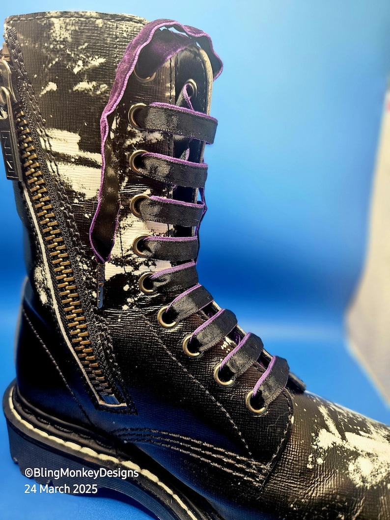 May include: Black leather boots with white paint splatter and purple laces. The boots have a side zipper and a white rubber sole.