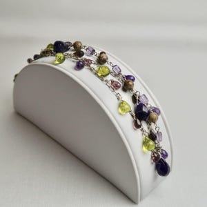 Può includere: Collana a più fili con catena argentata e una varietà di pietre preziose colorate. Le pietre includono perle viola, verdi, marroni e trasparenti. La collana è esposta su un supporto bianco curvo.