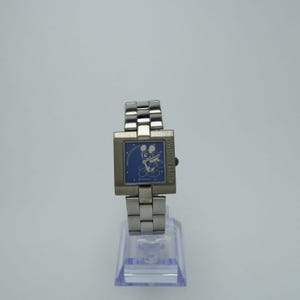 May include: A silver-tone wristwatch with a square case and a link bracelet. The blue watch face features a white graphic of Mickey Mouse playing a violin, the text "MILLENNIUM" on the right side, and "©DISNEY" at the bottom centre.
