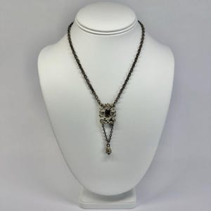 May include: A dark silver-toned necklace featuring a central rectangular pendant with an intricate, textured metal frame surrounding a dark stone. A delicate chain extends from the pendant, ending in a small, teardrop-shaped metal charm.