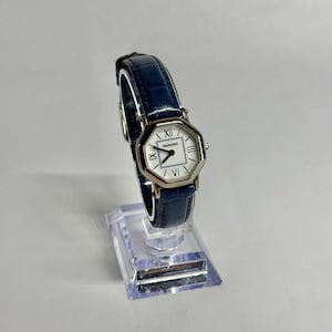 Könnte beinhalten: Eine Armbanduhr mit einem dunkelblauen, strukturierten Lederarmband und einem silberfarbenen achteckigen Gehäuse. Das weiße Zifferblatt zeigt schwarze römische Ziffern auf der Zwölf und Sechs, einfache Strichindizes und den Schriftzug marie claire.