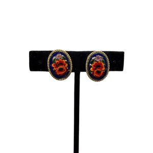 May include: Oval-shaped clip-on earrings with a floral design. The earrings feature a dark blue background with a gold-colored rope-like border. The floral design includes red, orange, and purple flowers.