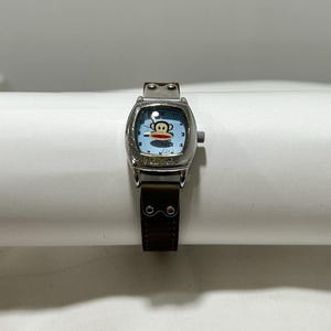 Könnte beinhalten: Eine silberne Armbanduhr mit rechteckigem Zifferblatt und braunem Lederarmband. Das Zifferblatt ist hellblau mit einer Cartoon-Affen-Grafik und den Worten "Paul Frank". Die Uhr hat silberne Stundenmarkierungen und Zeiger.