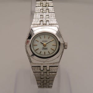 May include: A vintage Orient Charmak wristwatch with a silver metal strap and case. The watch face is white with gold hands and markers. The watch has a textured strap and a small crown. The watch face reads "Orient Charmak 21 Jewels."