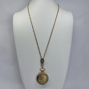 May include: A vintage gold-toned pocket watch pendant necklace. The round watch features an ornate design and is suspended from a long chain. The necklace is displayed on a white mannequin, showcasing its length and detail.