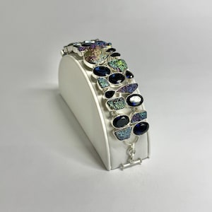 Può includere: Un bracciale in argento caratterizzato da una serie di pietre ovali blu scuro alternate a pietre druzy testurizzate e iridescenti. Le pietre sono incastonate in maglie di metallo argentato e il bracciale si chiude con una chiusura a T.