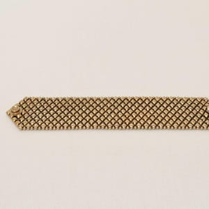 May include: A gold-toned bracelet with a mesh design. The bracelet is rectangular with rounded edges and features a series of small, closely-knit beads. The ends of the bracelet have a decorative button-like detail.