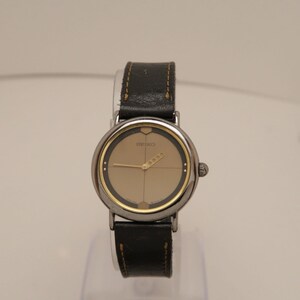 May include: A vintage Seiko watch with a silver case and a black leather strap. The watch face is a light tan colour with gold hands and markers. The word "SEIKO" is printed on the watch face.