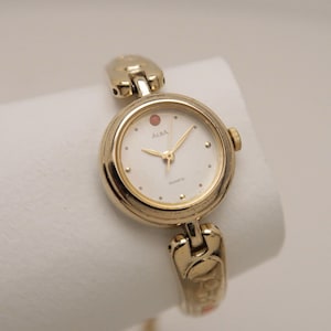 May include: Gold-toned ALBA quartz watch with a white face and gold hands. The watch has a round face with a small red accent and a gold band. The watch is displayed on a white surface.