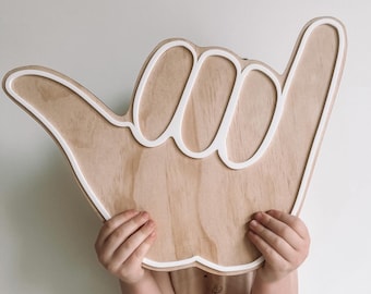 May include: A wooden hand-shaped sign with a white outline, shaped like a shaka hand gesture. The sign is being held in front of a child's face.