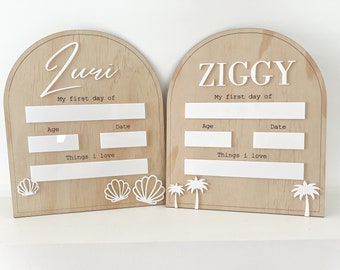 May include: Two wooden first day of school signs. One sign says "Juri" and the other says "Ziggy" in white lettering. Each sign has space for age, date, and things I love. The Juri sign has seashell cutouts, and the Ziggy sign has palm tree cutouts.