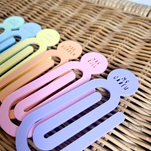 May include: A selection of pastel-coloured paper clips. Each clip features a circular top with text and a curved, elongated body. The colours are blue, yellow, orange, pink, and purple. The clips are arranged on a woven surface.