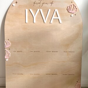 May include: Light brown wooden milestone board with an arched top. The board features the name "IYVA" in white letters, the text "first year of", and the months of the year. Pink seashell and star accents decorate the board.