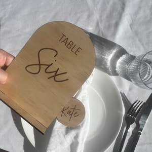 May include: A wooden table number six with the word "Table" engraved above the number. A smaller wooden circle with the name "Kate" is also visible. The table setting includes a white plate, cutlery, and a glass.